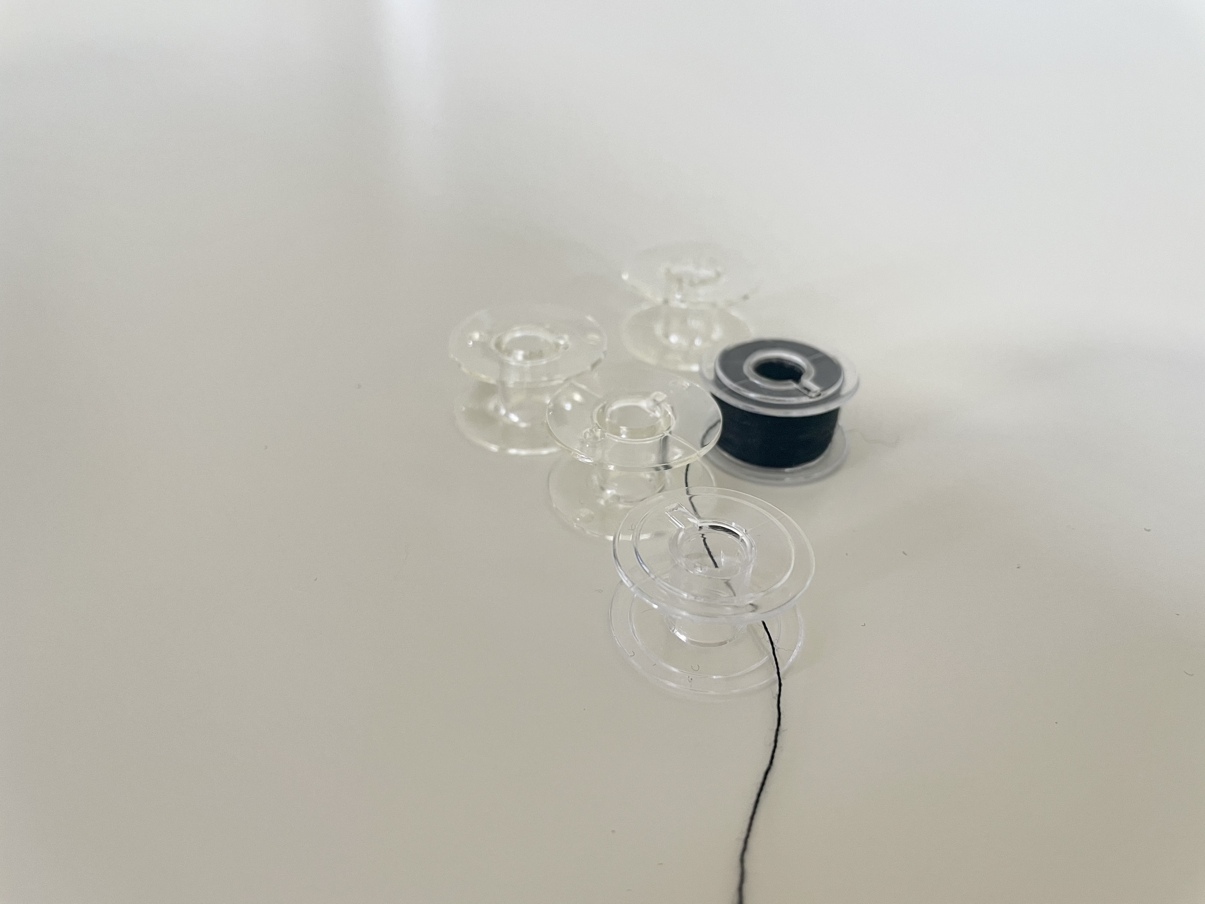 Five clear bobbins, one wound with black thread, sit on a white surface.