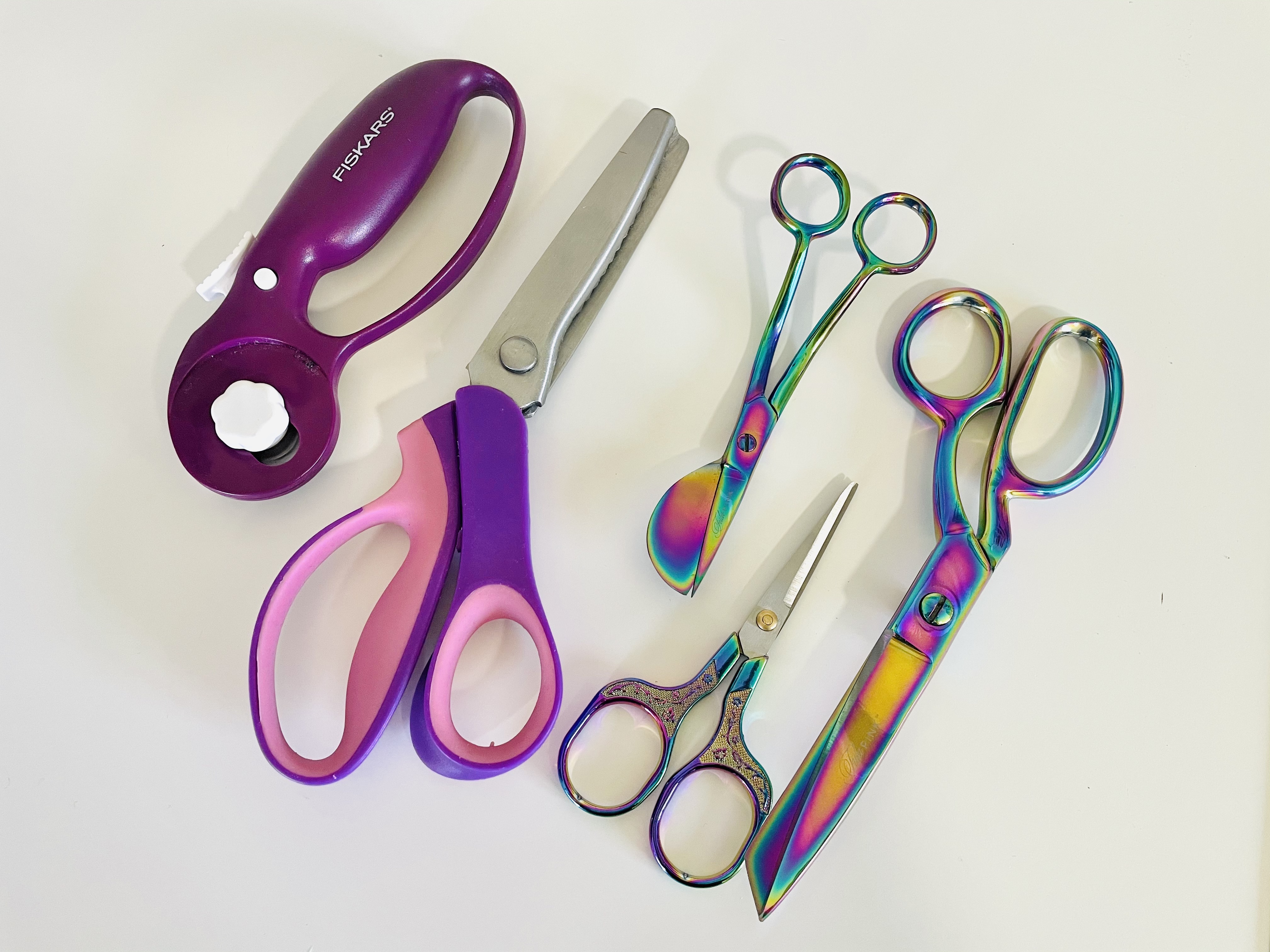 A purple rotary cutter, purple and pink pinking shears, oil slick rainbow duck bill scissors, oil slick thread snips, and oil slick fabric shears sit on a white backdrop