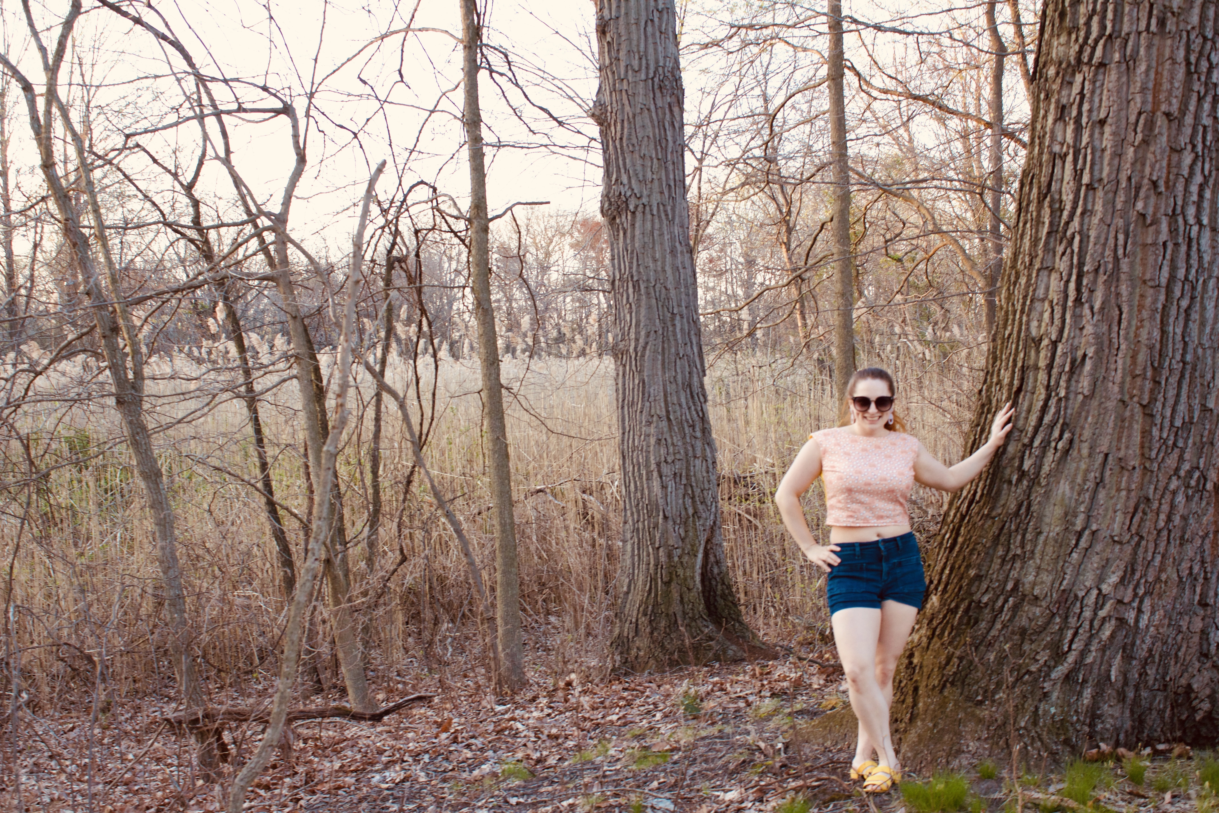 A person leans against a tree while wearing a pink crop top, and denim shorts.