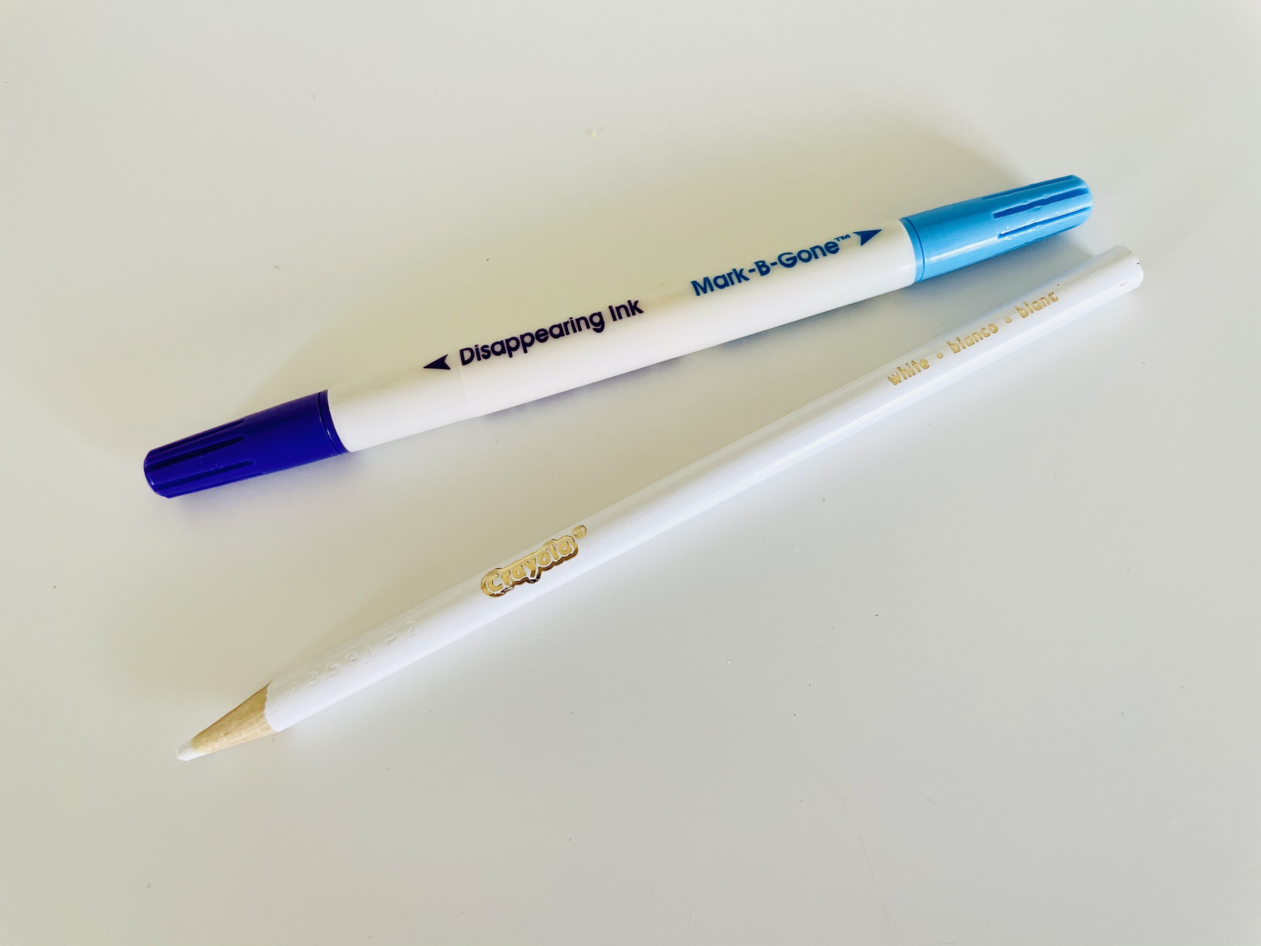 A double-sided Dritz Mark-be-Gone disappearing ink pen sits next to a Crayola white colored pencil on a white surface.