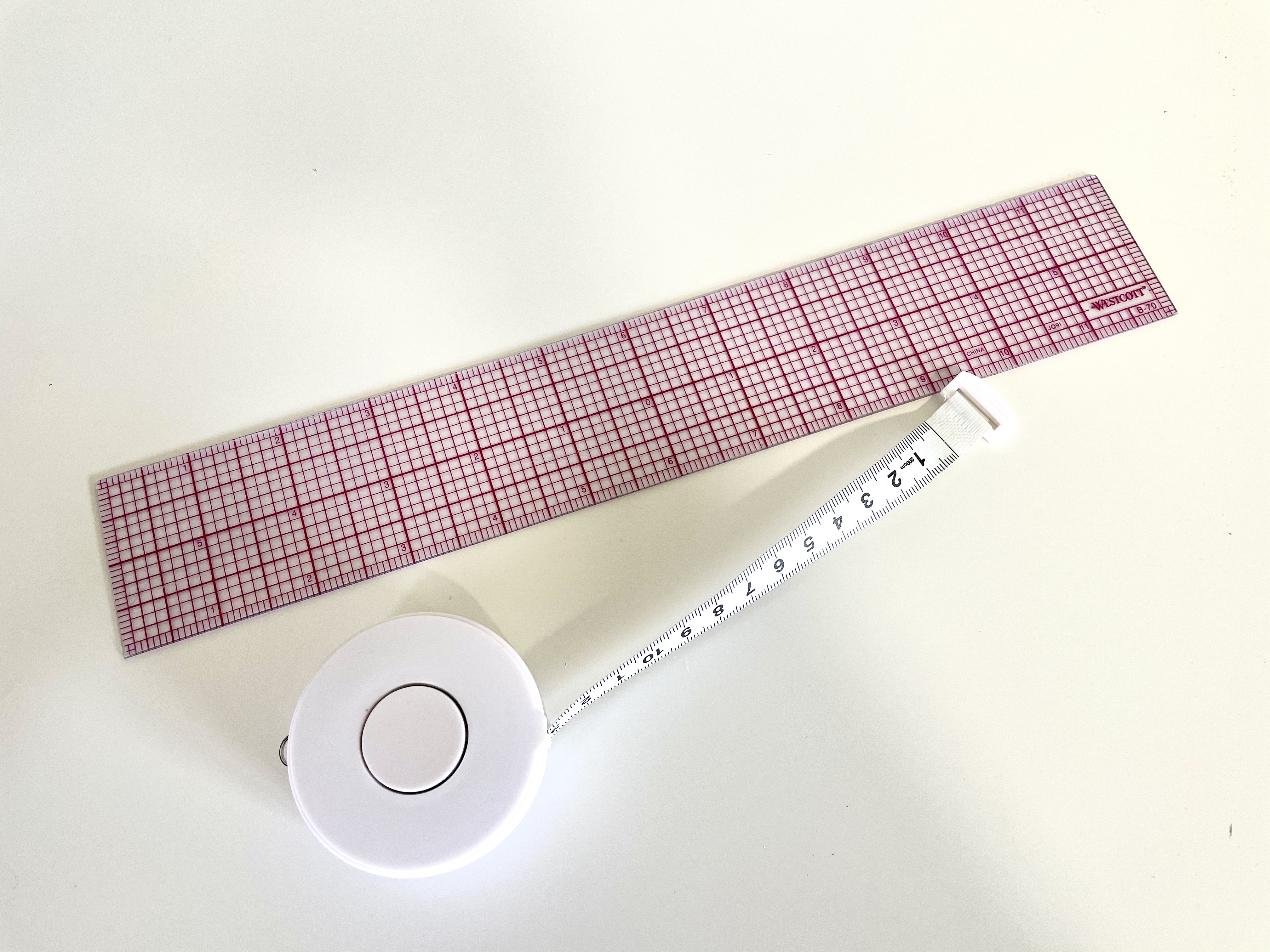 A quilter's ruler sits next to a retractable measuring tape on a white surface