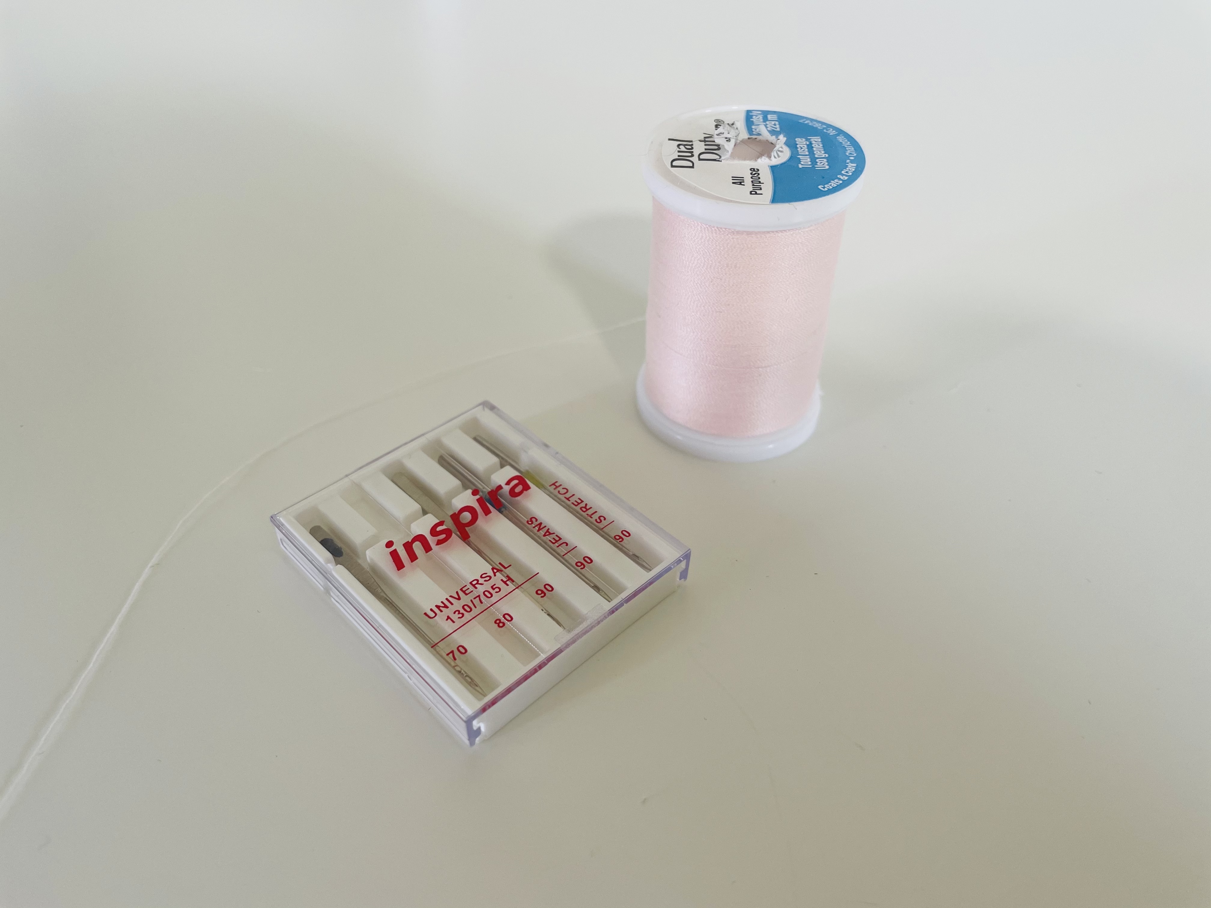 A spool of light pink Coats & Clark Dual Duty thread sits next to a package of various machine sewing needles