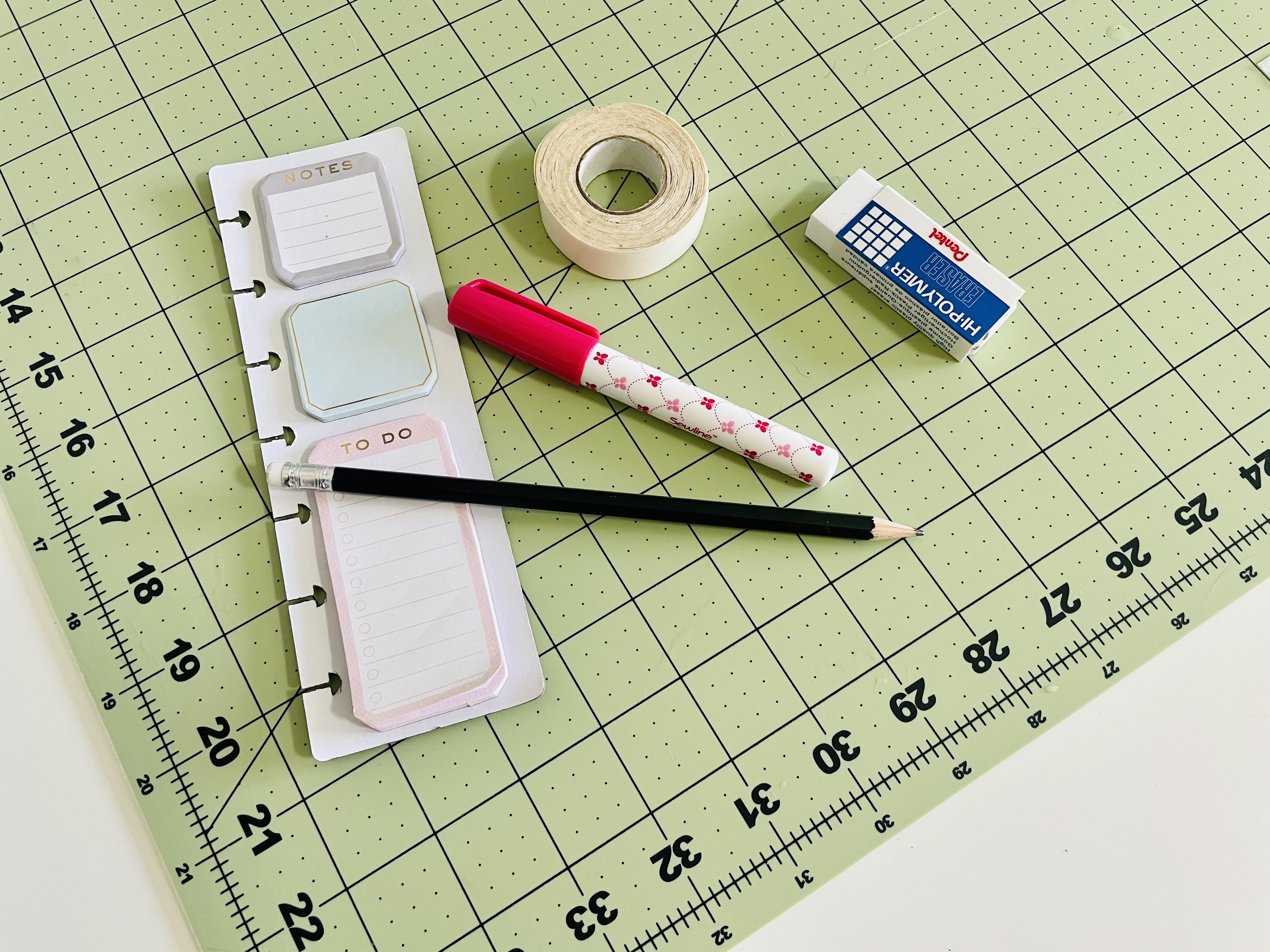 A pencil, sticky notes, fabric glue stick, fabric tape, and eraser sit on a green cutting mat