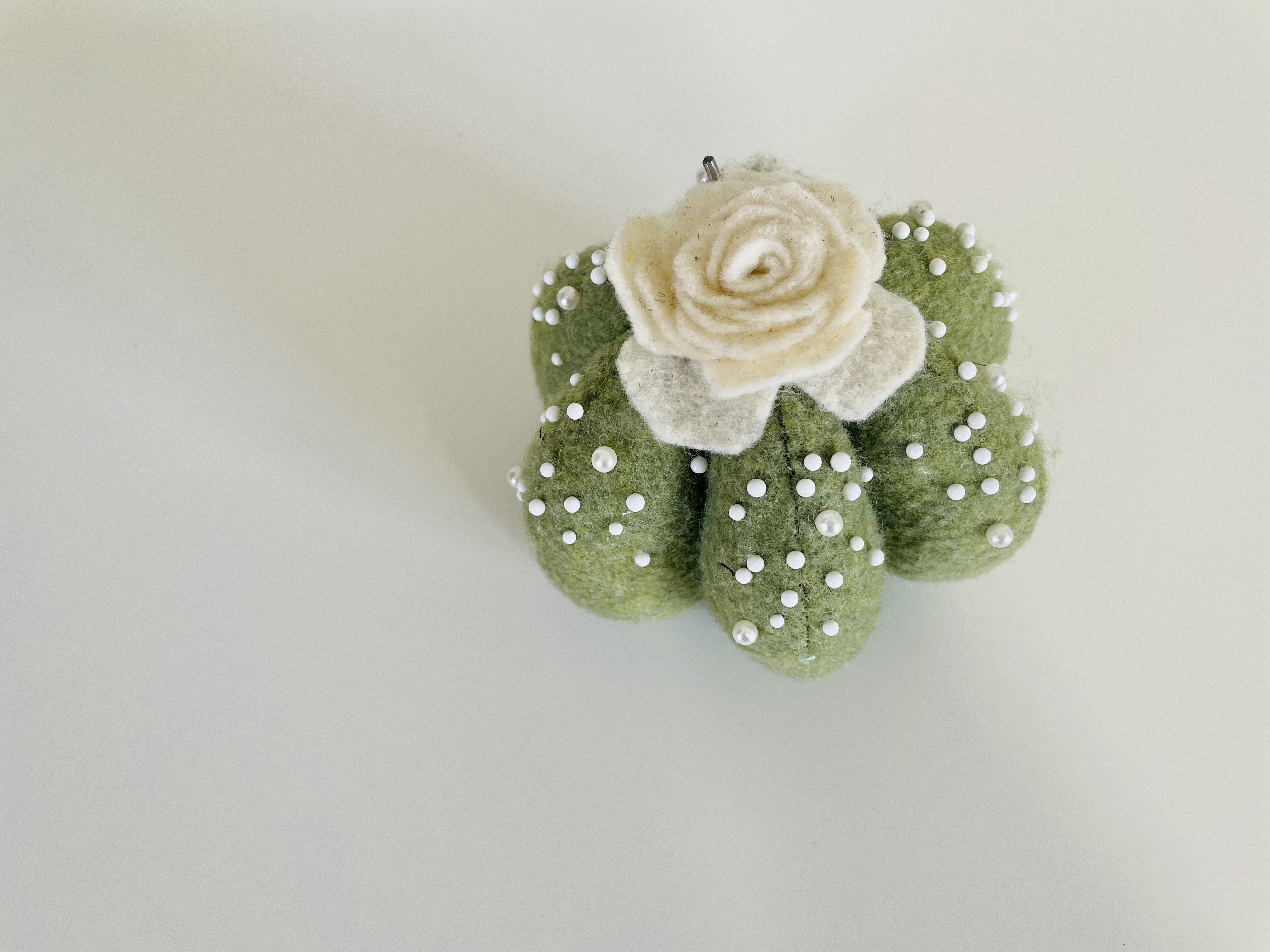 A succulent-shaped green pin cushion with a cream-colored flower and white pins throughout sits on a white background.