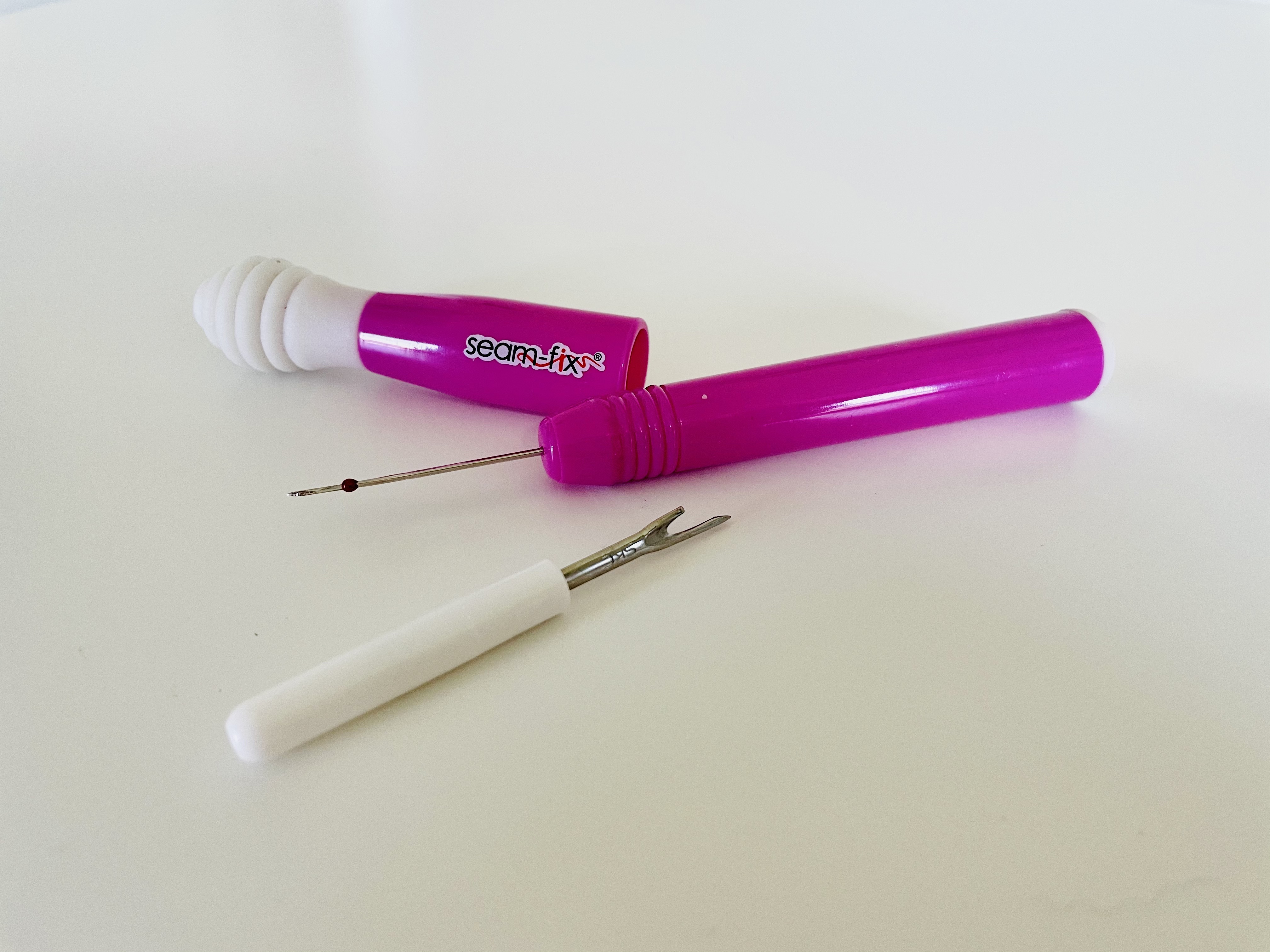 A hot large pink seam ripper sits next to a small white seam ripper on a white surface