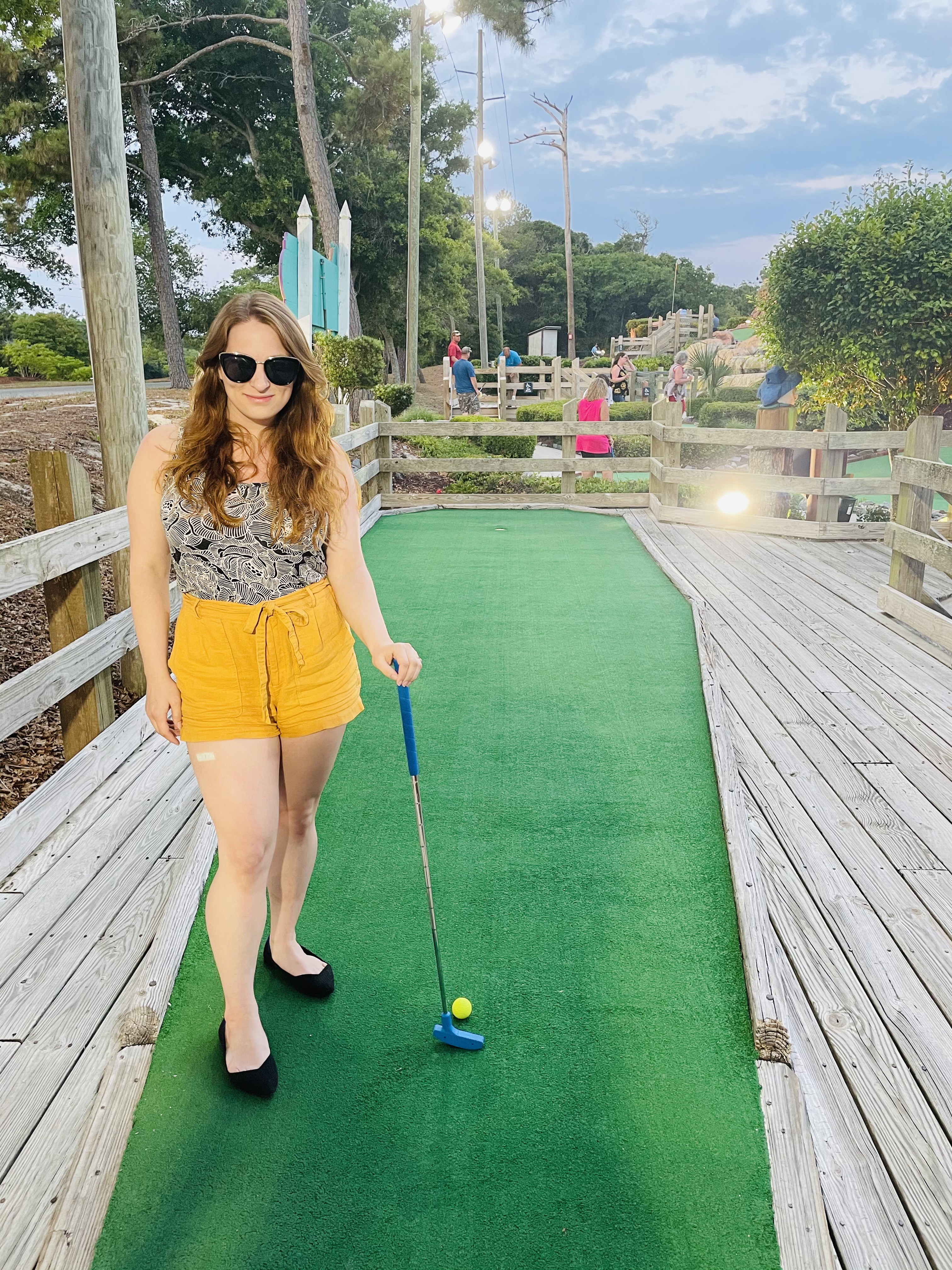 Lindsie wears her lago tank while standing on the green at a putt-putt mini golf course. She is holding a putter in her left hand and is wearing mustard shorts and sunglasses.