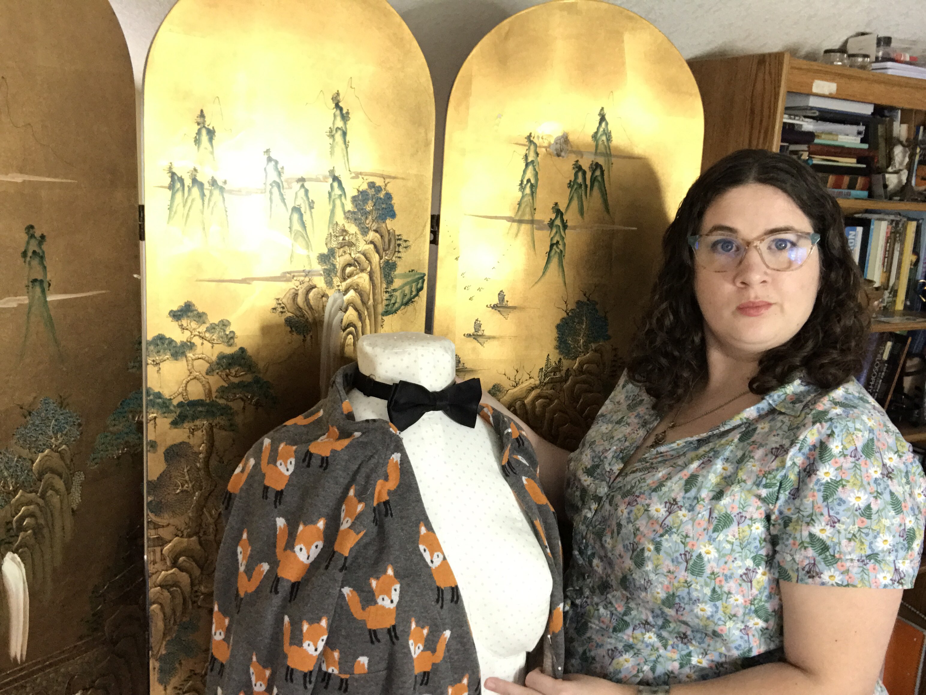 Jasmin stands next to a dress form, which has a gray fabric with orange foxes draped over it. The dress form is also wearing a black bow tie.