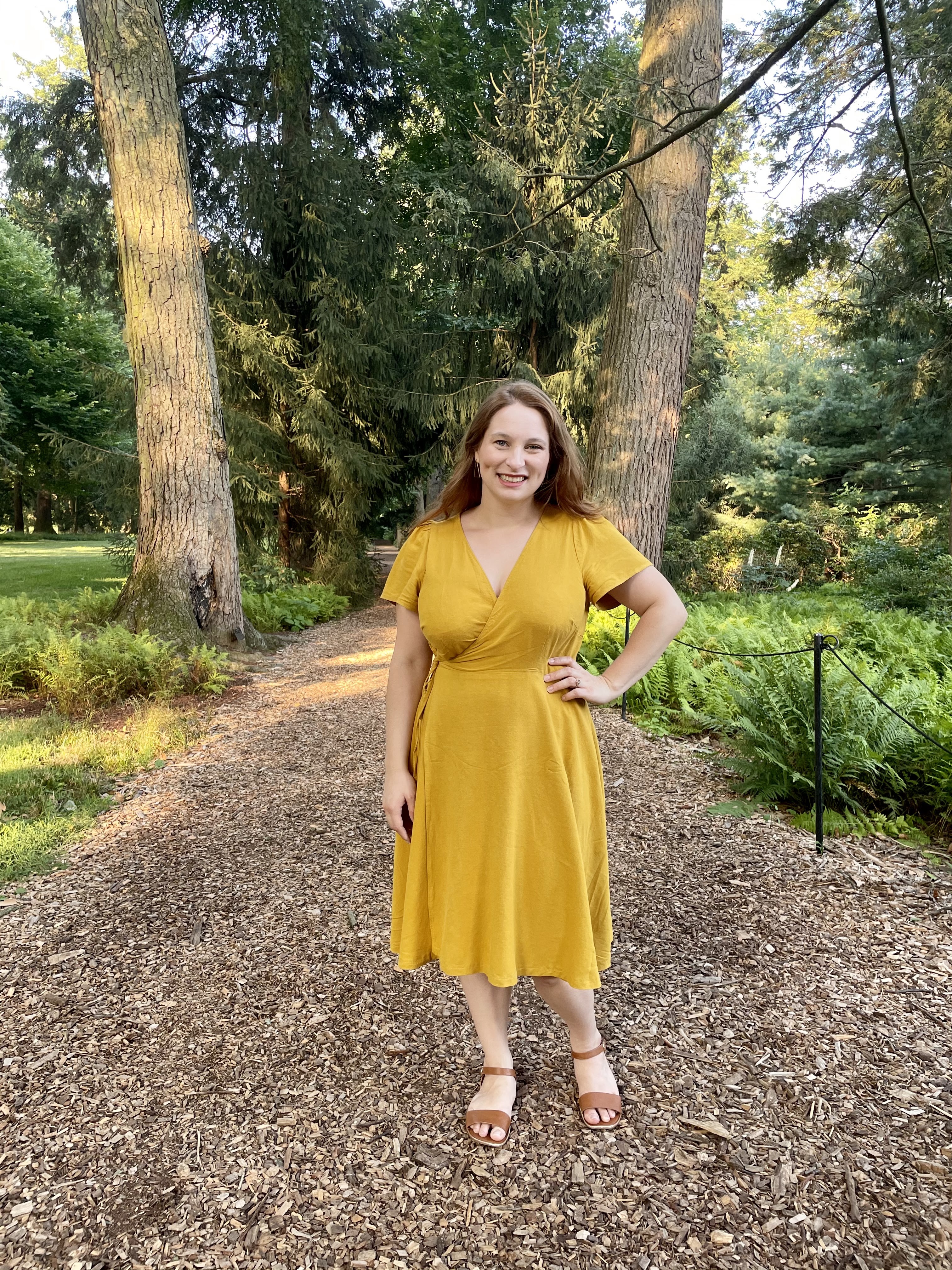 Lindsie wears her yellow Roseclair wrap dress in the woods. Lindsie is a white woman with large boobs, and she has paired her mustard yellow wrap dress with a pair of brown sandals. She stands on a bark trail with trees in the background.