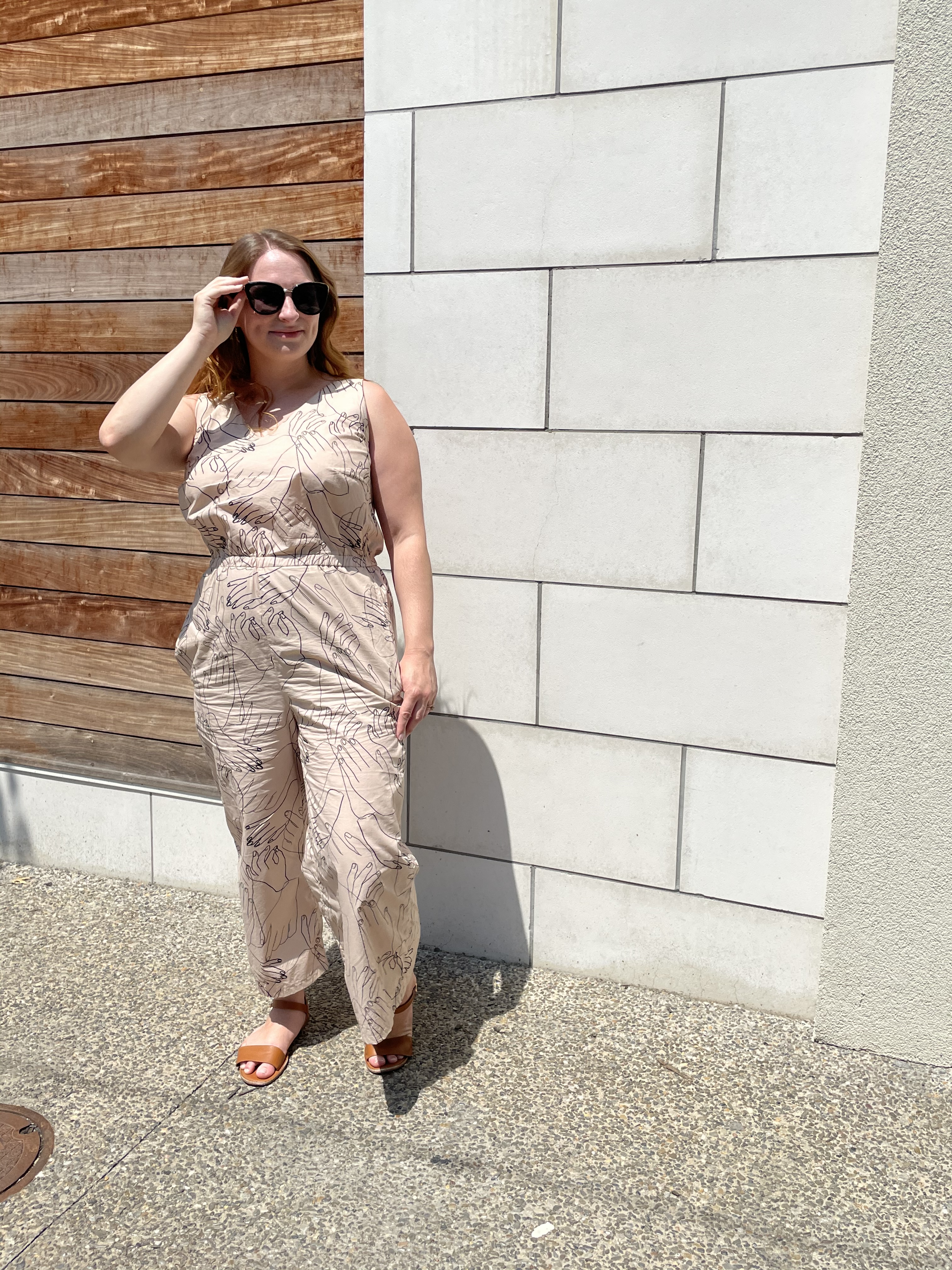 Lindsie, a white woman in her 20s, stands on a sidewalk in front of a wall that is partially wood and partially marble tiled and models a jumpsuit. The jumpsuit is sleeveless and 7/8 length and is made from a tan-colored material with black outlines of hands throughout. Lindsie has her hand on her sunglasses and looks in the distance. End.