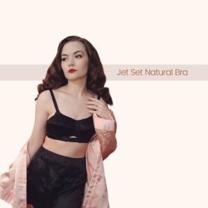 A front view of a model wearing the Jet Set Natural Bra Pattern in black satin with black elastic, a black satin half slip and a light pink robe with the text “Jet Set Natural Bra”.