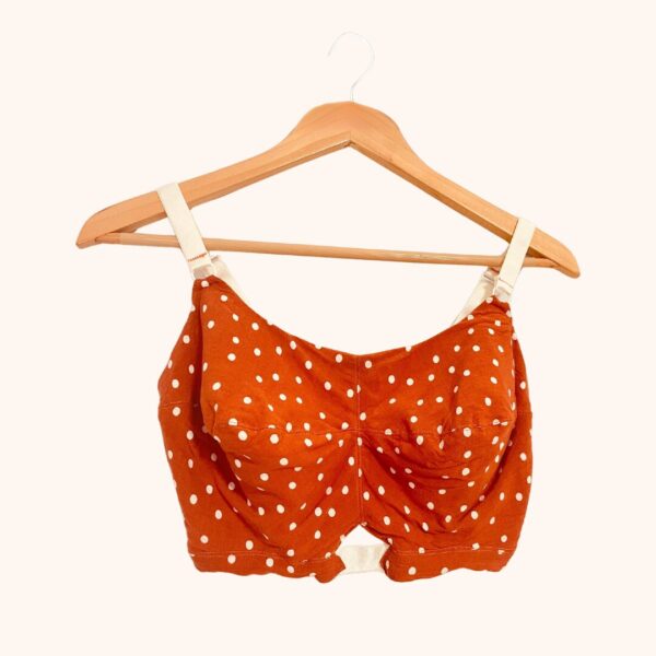 A front view of the Jet Set Natural Bra Pattern in an ivory polka dot pattern on tangerine fabric with ivory elastic on a wooden hanger.